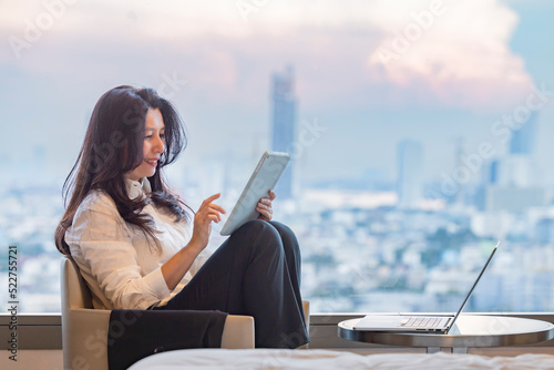 Asian woman use modern technology tablet and laptop, browsing internet, video call meeting with coworkers, checking email at home in the high floor of the building overlooking the city at sunrise time