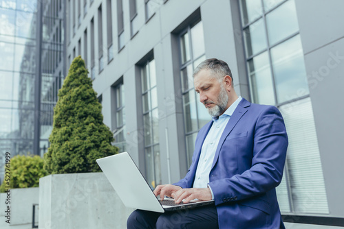 Serious and focused senior gray haired businessman outside office building, working using laptop, male banker investor in business clothes sitting on bench during break
