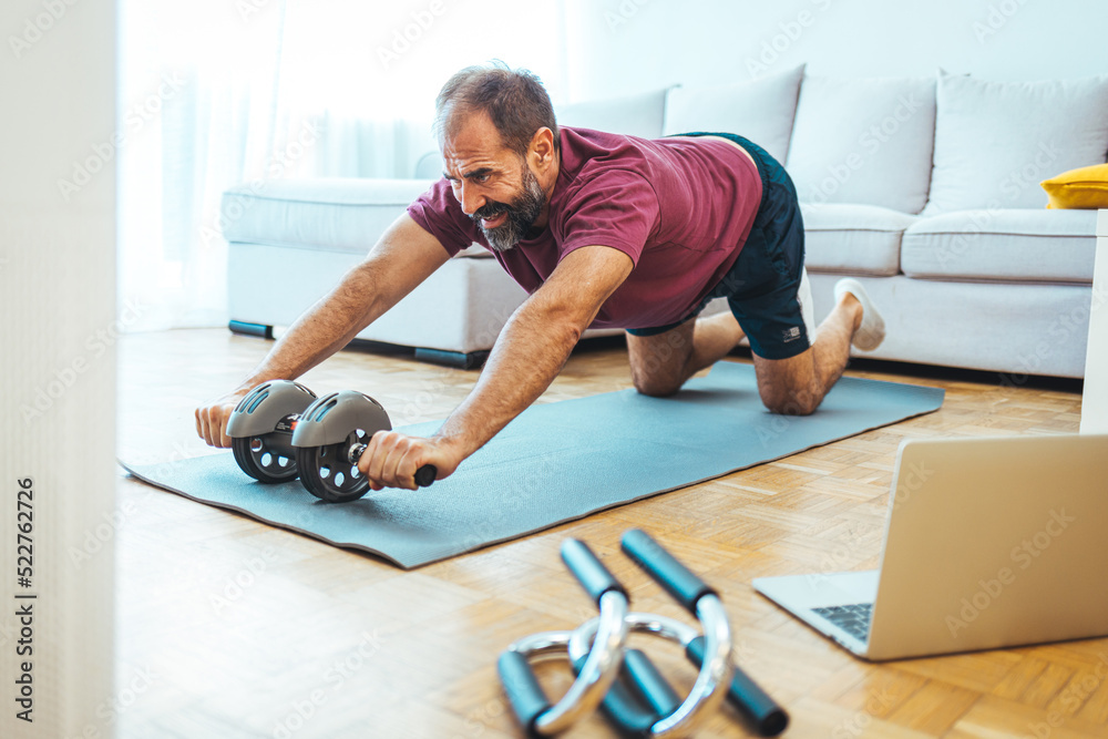 Mature Adult Man Exercising at Home in His Living Room. Mature man exercising at home - watching virtual exercise class on laptop. . Athletic man in sports clothes having domestic training Mature Adult Man Exercising at Home in His Living Room. Mature man exercising at home - watching virtual exercise class on laptop. . Athletic man in sports clothes having domestic training