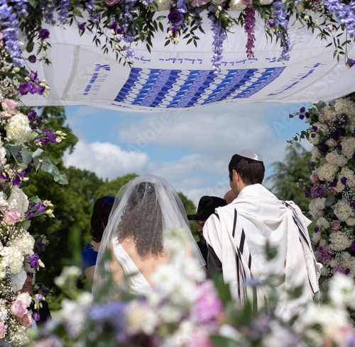 Jewish couple at a marriage ceremony under a floral chuppa wedding canopy. Canopy is inscribed in Hebrew with words from the biblical book Song of Songs: 'I am to my beloved'.