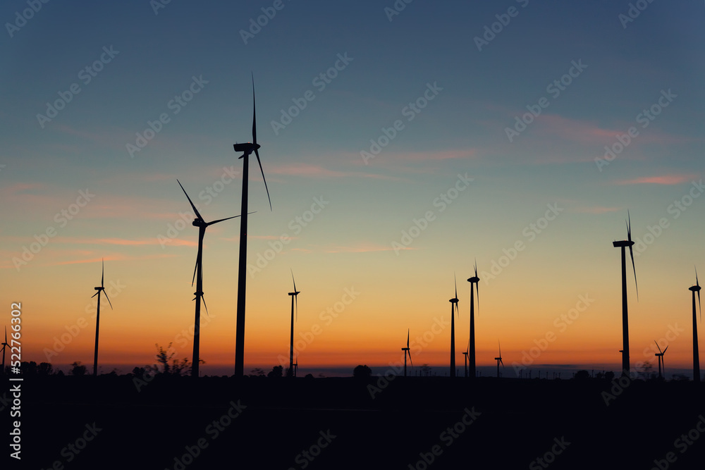 Fototapeta premium Panoramic scenic landscape view new modern wind turbine farm power generation station against fiery warm sunset sky field. Clean sustainable zero emission alternative electricity windfarm industry