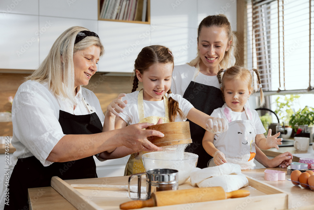 Young mom daughters and grandmother spending time in the kitchen baking cooking cakes cookies pie. Granny teaching grandchldren sifting flour through sieve. Women having fun together. Young mom daughters and grandmother spending time in the kitchen baking cooking cakes cookies pie. Granny teaching grandchldren sifting flour through sieve. Women having fun together.