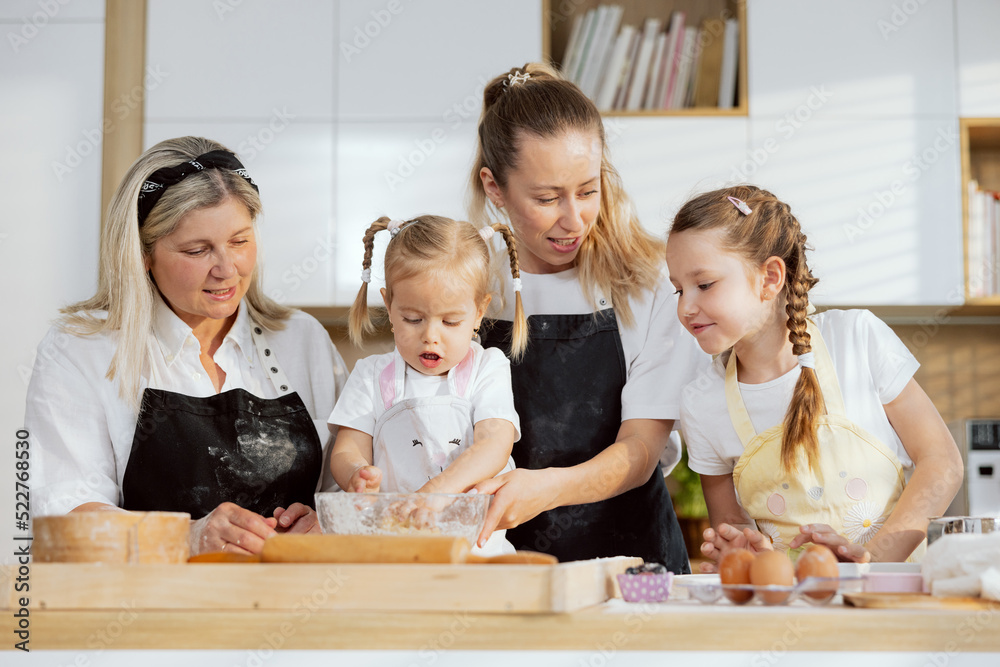Excited family teaching girls kneading dough standing at table in modern light kitchen. Preschooler daughter helping holding knife cutting butter with mother help. Granny managing process. Excited family teaching girls kneading dough standing at table in modern light kitchen. Preschooler daughter helping holding knife cutting butter with mother help. Granny managing process.