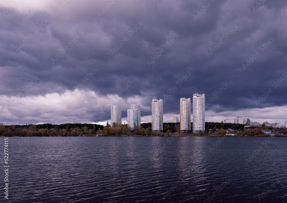Fototapeta premium View across the dark lake to tall new houses in the city near nature. Fantasy nature. Dark cloudy landscape. Dark terrible clouds