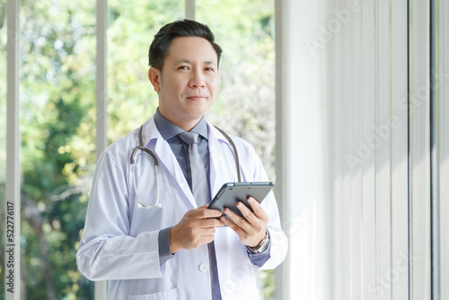 Portrait of asian senior doctor standing in medical office while using tablet.