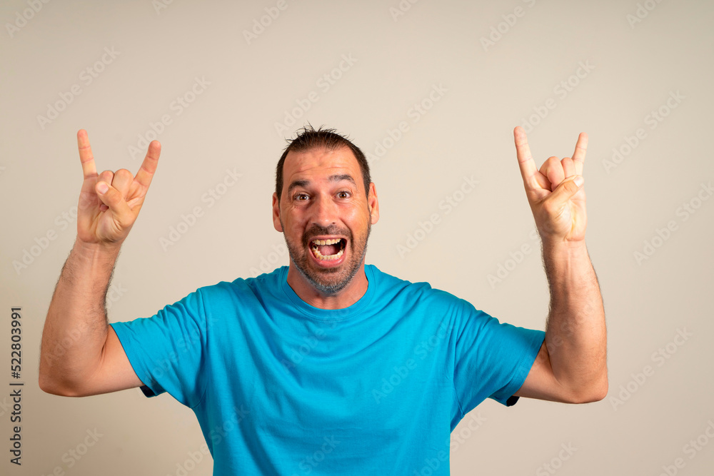 Rock fan gestures. Caucasian bearded man in blue t-shirt making rock symbol with hands up while standing against beige background. body language concept. Rock fan gestures. Caucasian bearded man in blue t-shirt making rock symbol with hands up while standing against beige background. body language concept.