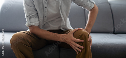 Young man who has hurt his knee is sitting on sofa and hugging his leg with grimace of suffering on face. Unhappy guy with rheumatism, arthritis or osteoarthritis has intense severe pain in knee cap