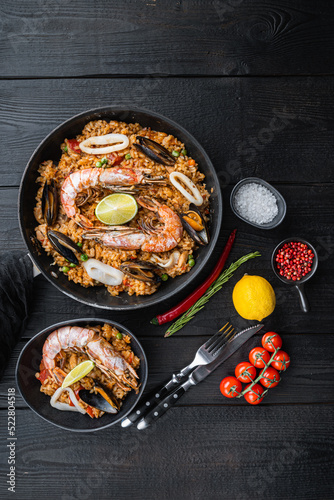 Traditional spanish seafood paella in pan and bowl with rice, peas, shrimps, mussels and squid on black wooden planks, top view