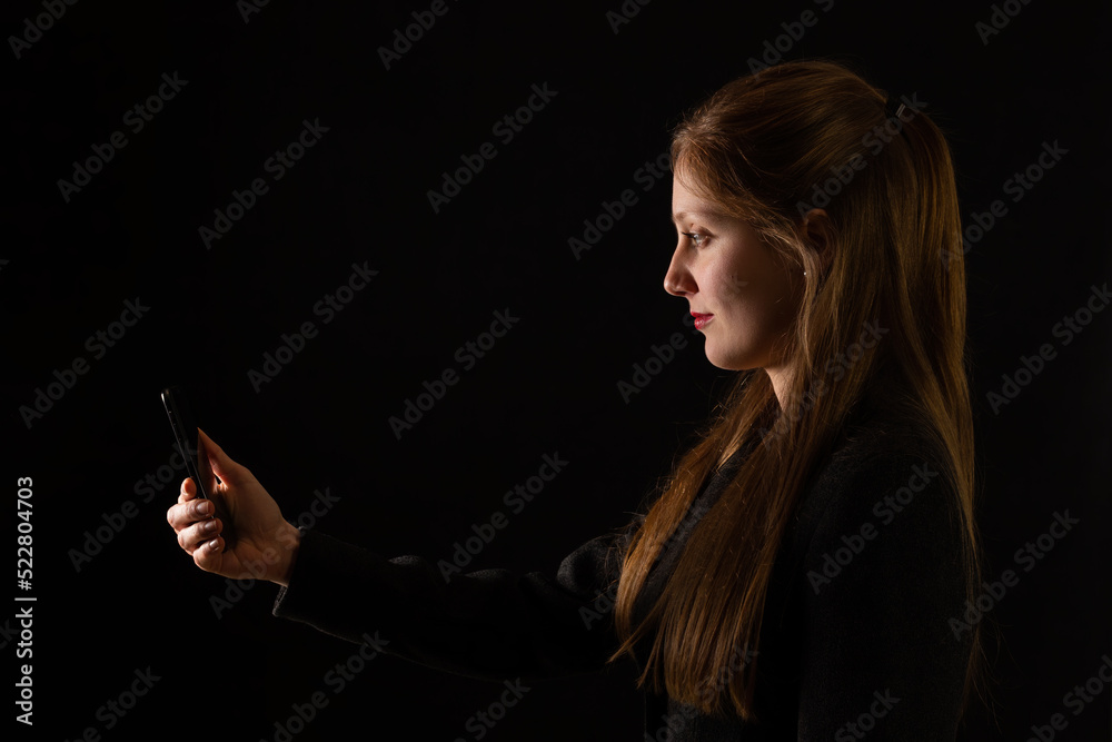 Young Caucasian woman holding smartphone in front of her face with black background. Side view portrait of female office worker interacting with modern technology at work.