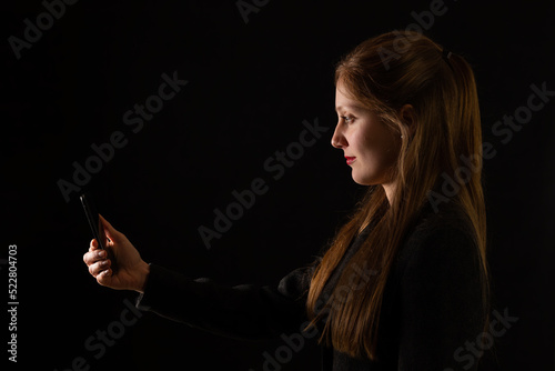 Obraz na płótnie Young Caucasian woman holding smartphone in front of her face with black background. Side view portrait of female office worker interacting with modern technology at work. w salonie