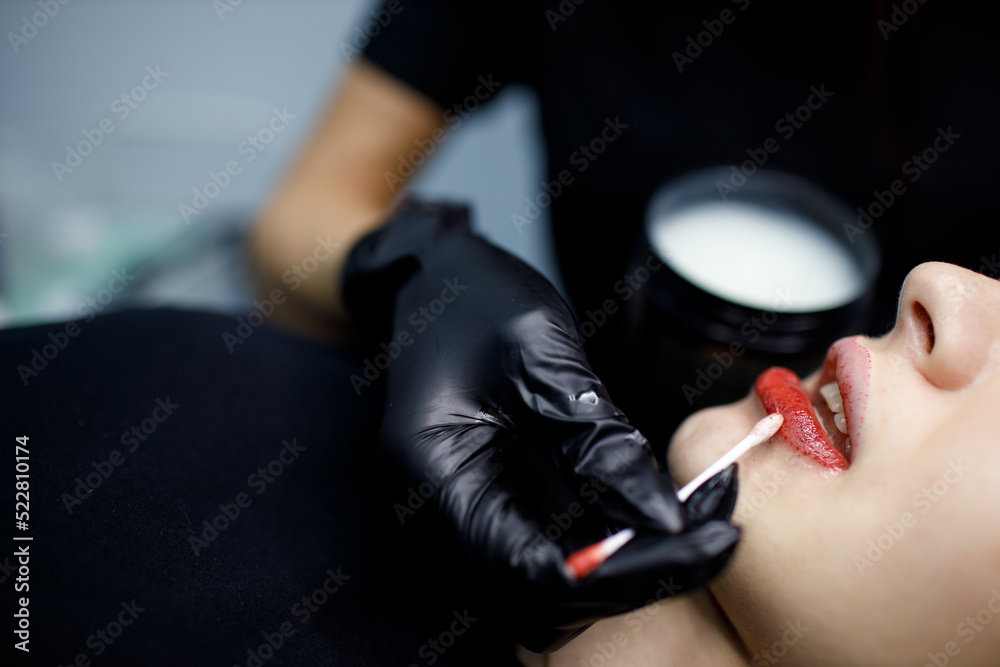 Cosmetologist applying vaseline petrolatum to permanent make up tattoo on young woman lips. Permanent lips tattoo procedure close up