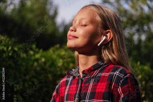 Smiling little girl listening music in earphones standing with closed eyes outdoor. Kid enjoying music