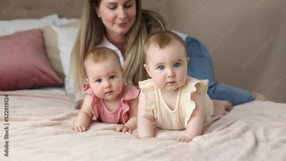 A young mother happy with two little siblings daughters, plays and cares for cute sweet twins 6 months old on a bed in a cozy home A young mother happy with two little siblings daughters, plays and cares for cute sweet twins 6 months old on a bed in a cozy home