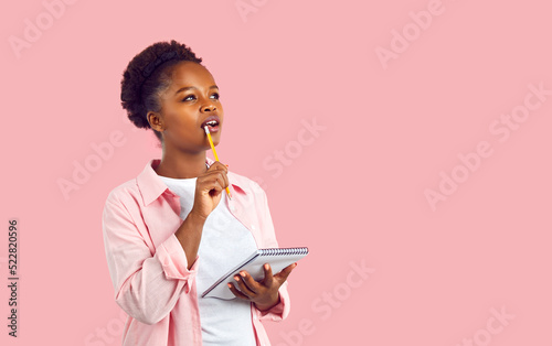 Beautiful Afro American woman in casual shirt standing on blank pink background, holding notebook, looking away, biting pencil and thinking. Female student or copywriter writing ideas for new article