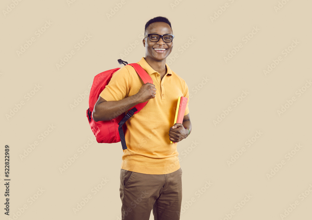Studio portrait of happy school, college or university student. Handsome young African American man in polo shirt and glasses standing on beige background, holding books and backpack and smiling Studio portrait of happy school, college or university student. Handsome young African American man in polo shirt and glasses standing on beige background, holding books and backpack and smiling