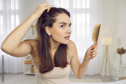 Portrait of woman in her 20s or 30s looking at her reflection with scared nervous expression as she notices bad signs like scalp dandruff, hair thinning, or hair falling out. Hair loss problem concept
