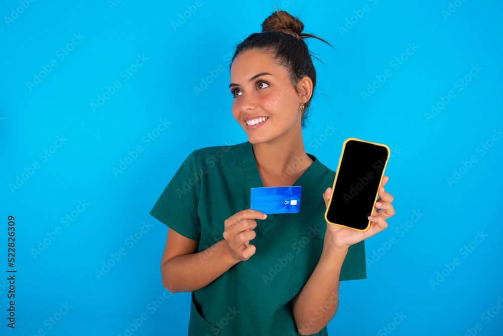 beautiful doctor woman wearing medical uniform over blue background holding bank card modern device looking empty space beautiful doctor woman wearing medical uniform over blue background holding bank card modern device looking empty space