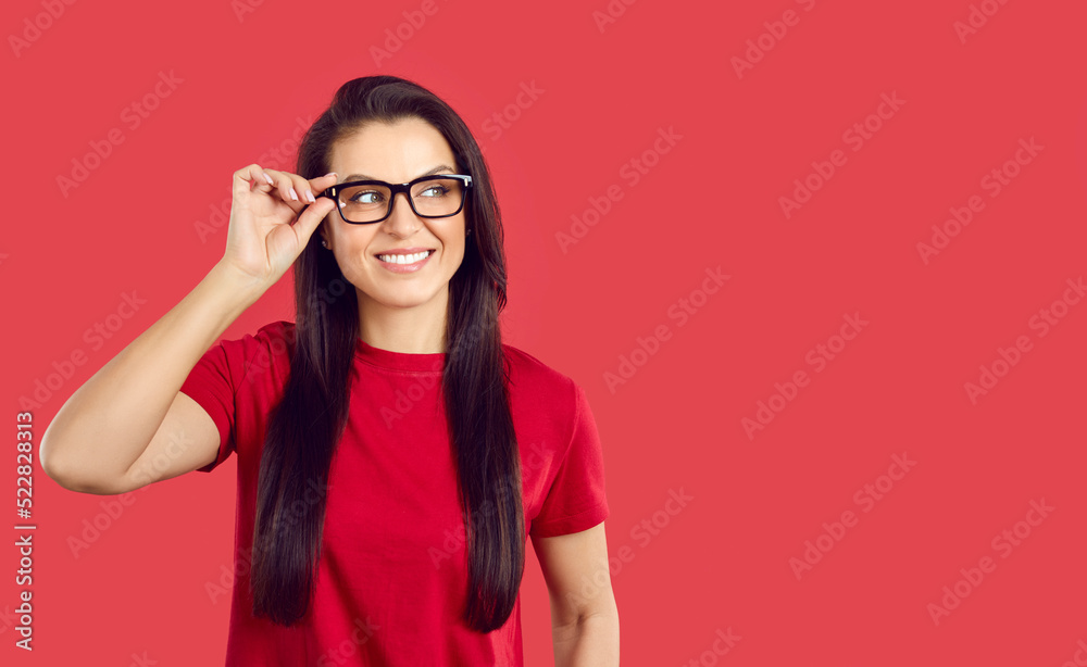 Portrait of happy millennial woman in classic eyeglasses with black frame isolated on red background. Joyful smart and confident casual woman smiling looking at copy space adjusting her glasses.
