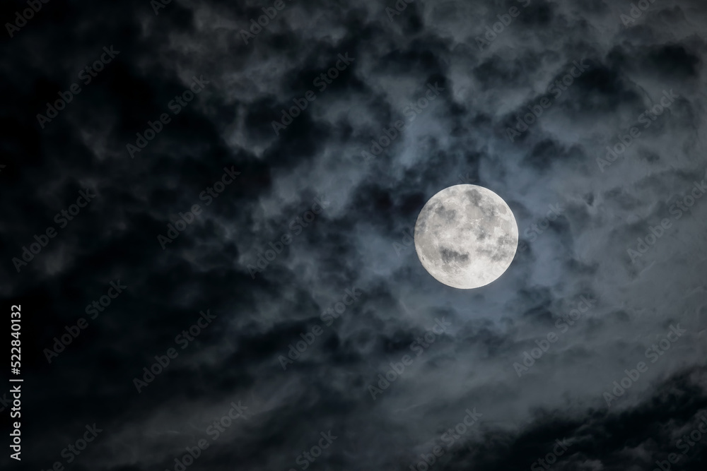 Full moon at night. The illuminated face of the moon is wrapped in a cloud cover that covers it in a veiled way. Full moon at night. The illuminated face of the moon is wrapped in a cloud cover that covers it in a veiled way.