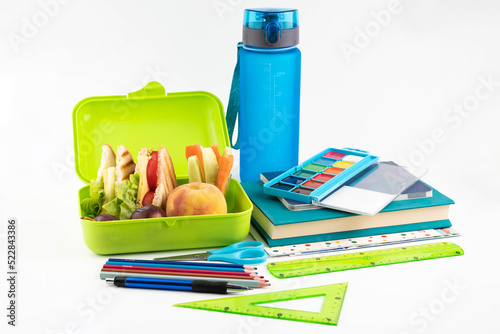 On a white background, a lunch box with a sandwich, vegetables and fruits, a bottle of water and school supplies.