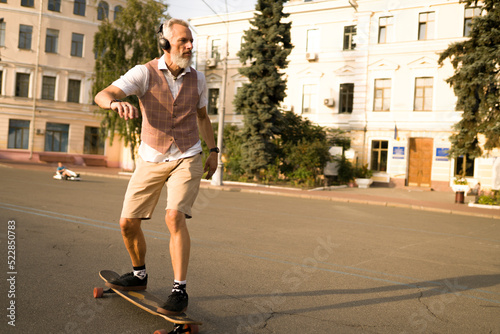 Portrait of handsome man in white shirt on sunset evening in summer. Stylish guy ride on skateboard on city street. Urban male lifestyle on buildings background