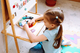 Top view of cute little girl sitting at white magnetic board with colorful numbers, playing wooden numbers on clock.