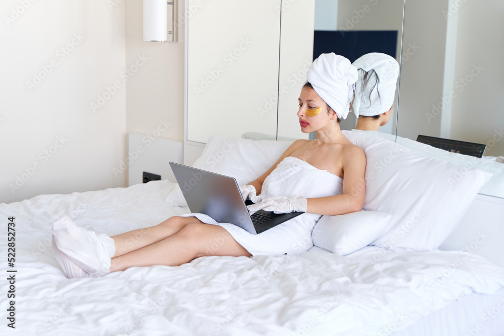 After a shower, a girl wrapped in a towel works on a laptop and uses cosmetic patches for skin under the eyes, lips and gloves to moisturize her hands and feet. Cosmetic trends for body care at home After a shower, a girl wrapped in a towel works on a laptop and uses cosmetic patches for skin under the eyes, lips and gloves to moisturize her hands and feet. Cosmetic trends for body care at home