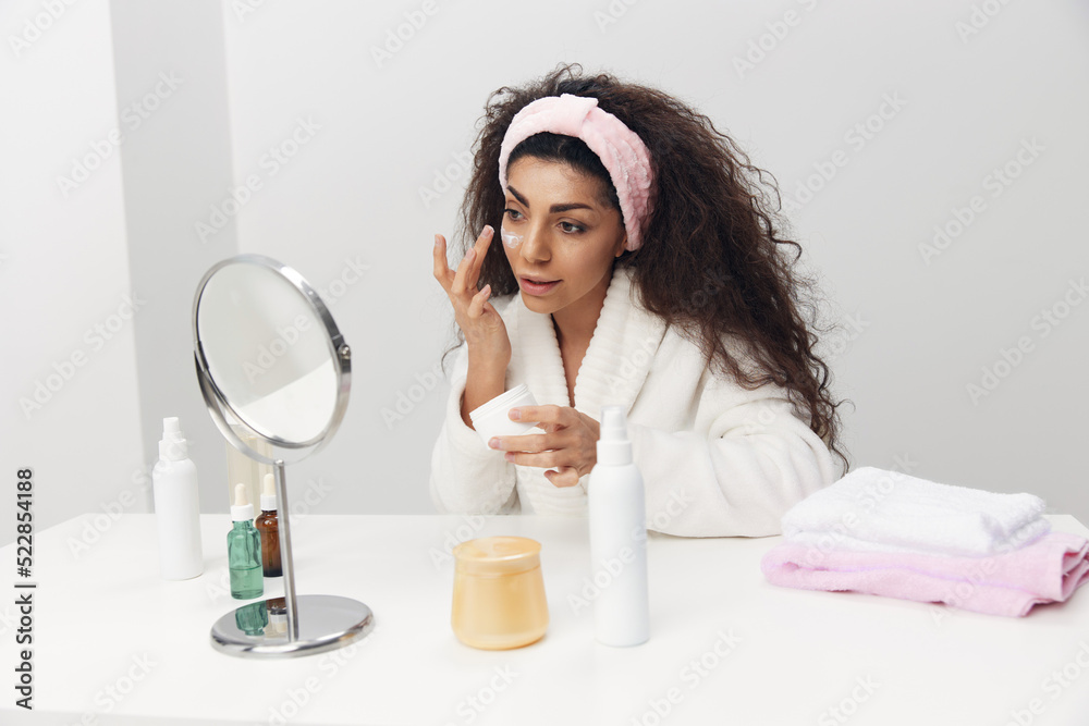 Cute tanned pretty curly Latin lady apply facial cream at cheek admiring herself in mirror doing beauty procedure in home interior. Copy space. Aesthetic medicine skincare beauty products ad concept Cute tanned pretty curly Latin lady apply facial cream at cheek admiring herself in mirror doing beauty procedure in home interior. Copy space. Aesthetic medicine skincare beauty products ad concept