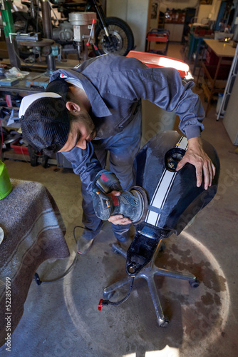 Full body of professional male mechanic in workwear polishing motorcycle fuel tank with grinding machine while working in garage