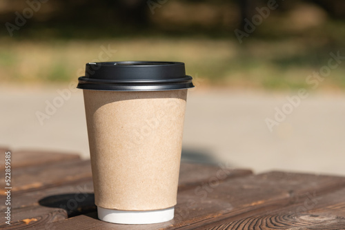 A paper cup of coffee with a lid stands on a rustic bench table against the backdrop of green foliage and sandy beach, sunny morning, breakfast, coffee break, coffee to go - blurred bokeh background