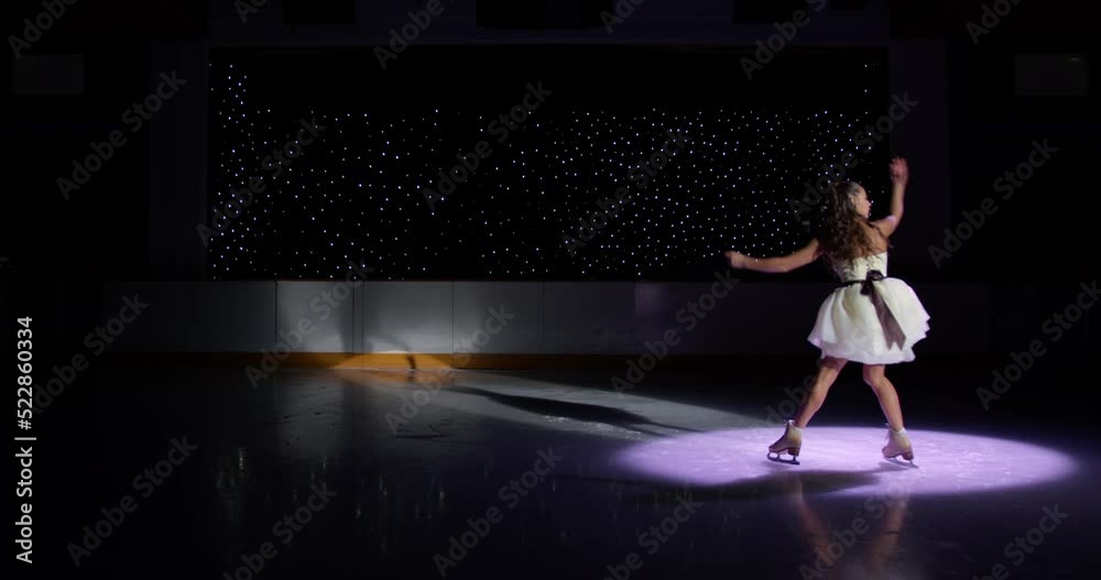 Beautiful ice skater girl in white dress dances on dark ice rink in spotlight, figure skating performance