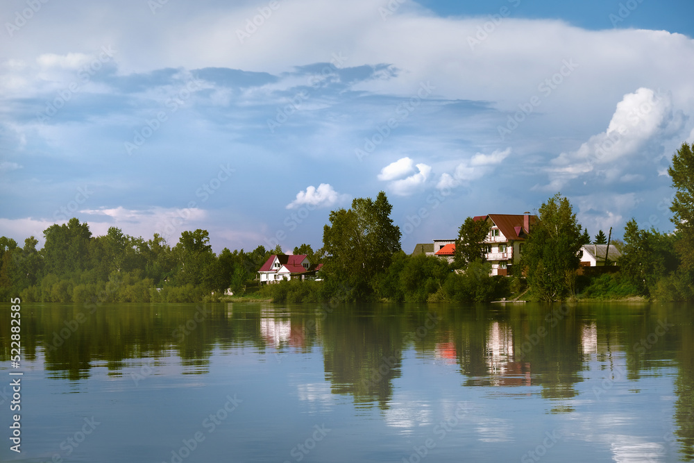 Obraz premium Summer landscape with river, along the banks of which wooden houses are nestled in lush greenery. Forest trees and sky are seen as a reflection on the water of a pond. Idyllic landscape