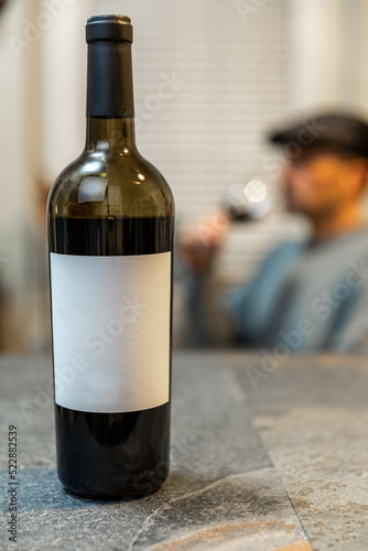 wine bottle with blank white label and person in background drinking from wine glass