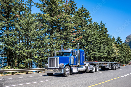 Blue classic big rig semi truck tractor with extended cab transporting empty flat bed semi trailer driving on wide highway road with trees on the side