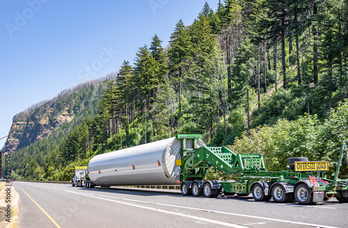 Heavy duty big rig semi truck transporting part of long oversized windmill generator pole on the specialized semi trailer with additional trolleys and oversize load sign on the back