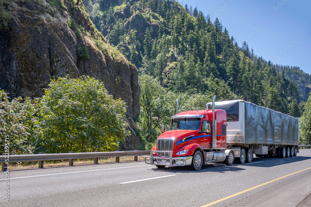 Low cab profile bright red big rig semi truck transporting cargo in covered framed semi trailer moving on the wide highway road with forest and rocky mountain on the side