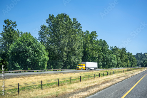 Low cab profile yellow big rig bonnet semi truck transporting cargo in dry van semi trailer driving on the highway road with grass divider line
