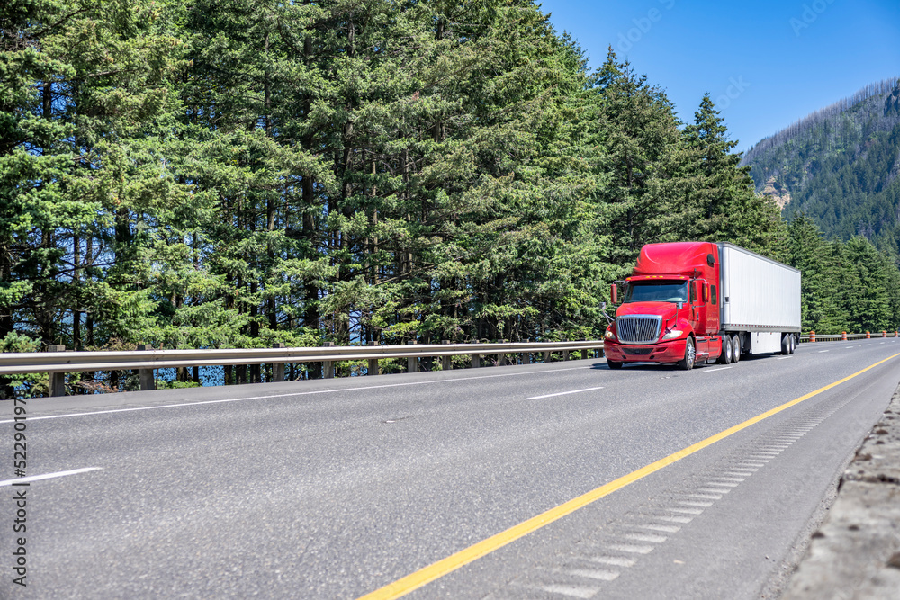 Red big rig semi truck with truck driver sleeping compartment transporting cargo in dry van semi trailer running long haul freight on the highway road with forest and mountain in Oregon