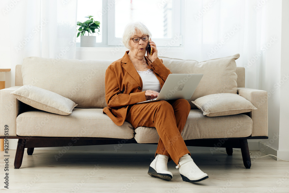 an emotional elderly woman with gray hair in a brown stylish suit and white boots is sitting on a beige sofa holding a laptop on her lap working from home an emotional elderly woman with gray hair in a brown stylish suit and white boots is sitting on a beige sofa holding a laptop on her lap working from home