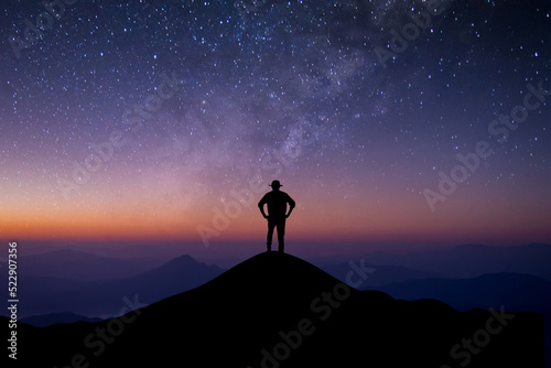 Silhouette of young man standing alone on top of mountain enjoying watching beautiful of night sky, star and milky way over the sky background. He was happy to travel alone.