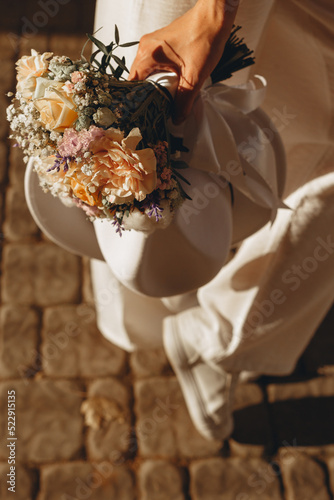 From above of crop bride in white sneakers and pants standing with hat and elegant wedding bouquet with tender pastel colored flowers at sunset 