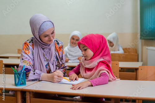 Female hijab Muslim teacher helps student girl to finish the lesson durind the school class in the classroom.	 photo