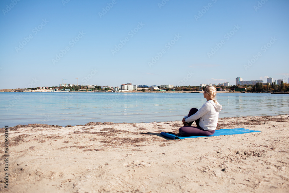 Elder female person takes care of body and does yoga by the sea outside in sunny weather. Female dressed the sport wear and do sport exercises in sandy beach