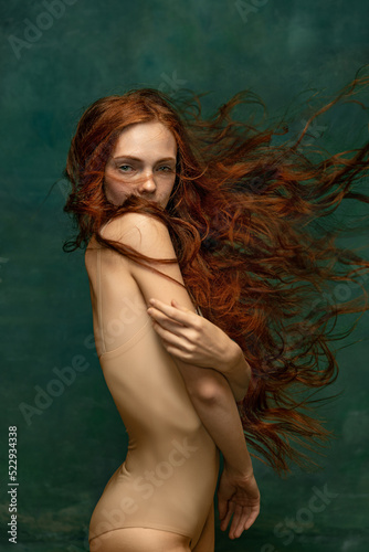 Young attractive redhead girl with long curly hair wearing nude color lingerie isolated over dark green background. Magic look. Concept of beauty, art photo