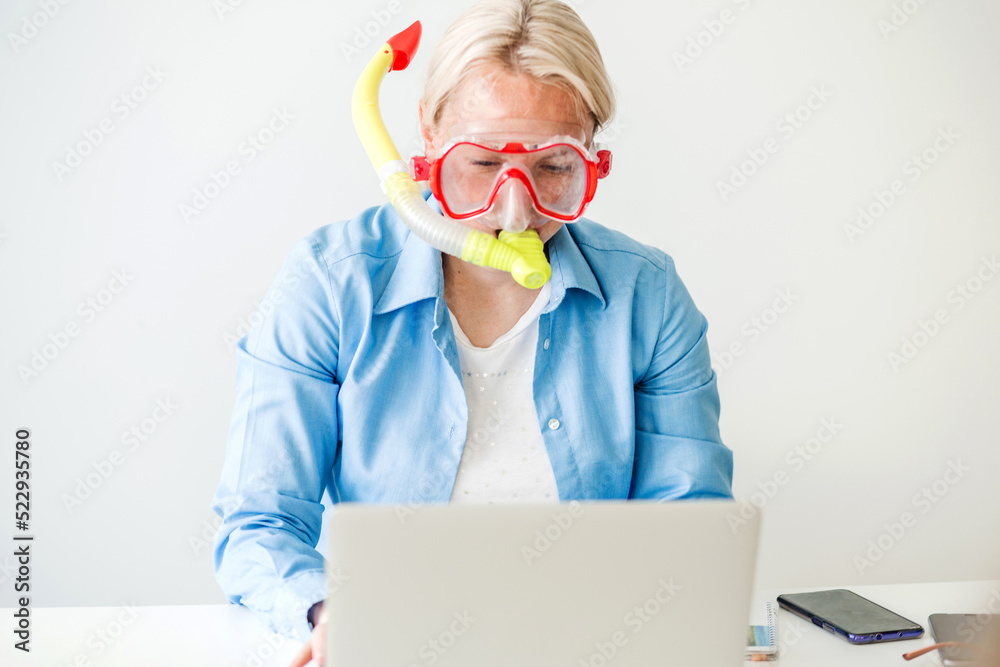 A blonde woman in a diving mask sits in the office, orders a tourist trip online, tickets for a plane, bus or train. Woman dreaming of sea at workplace A blonde woman in a diving mask sits in the office, orders a tourist trip online, tickets for a plane, bus or train. Woman dreaming of sea at workplace