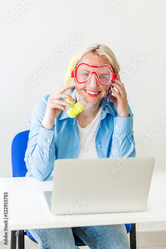 A blonde woman in a diving mask sits in the office and happily announces by phone that she is going on a trip or communicating with a tour operator