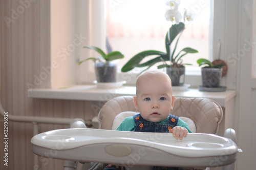 hungry boy sits waiting for food at an empty table for feeding in the kitchen on the windowsill of an orchid.