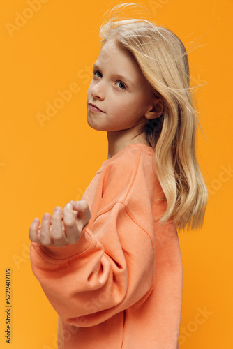 beautiful cute school age girl stands outstretching her hands to the camera on an orange background in bright clothes
