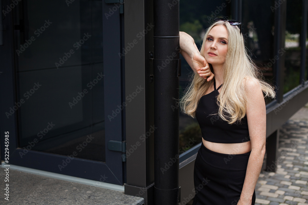 A stylish slender girl with blond long hair and a pleasant look stands against the backdrop of a modern building. Dressed in a short black sexy skirt and top. Sunglasses are on the head.