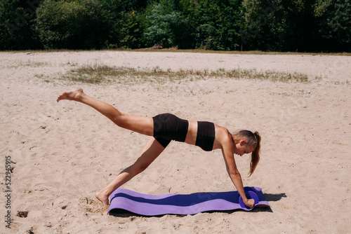 A beautiful tanned girl with a slim figure and blond hair, in black shorts and a tank top, is doing fitness with dumbbells, on a mat, on the beach in summer.